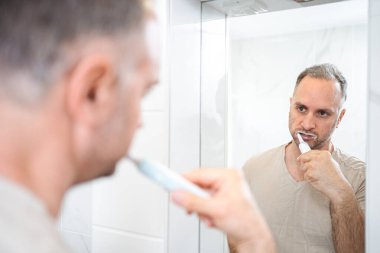 Adam banyoda dişlerini elektrikli diş fırçasıyla fırçalıyor..