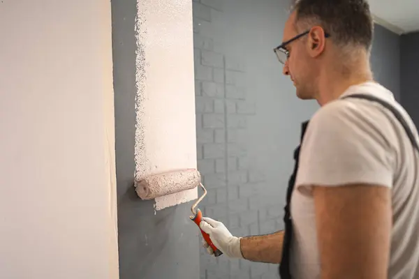 stock image A man is seen in the process of painting a wall with white paint. He holds a paint roller in his hand, carefully covering the surface with fresh paint. The wall is transforming from its previous color