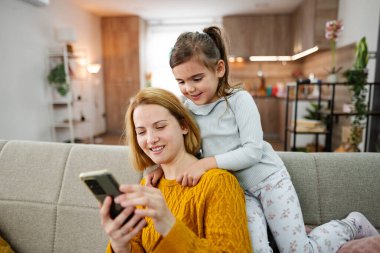 Anne ve kızı oturma odasında kanepede oturuyor, gülümsüyor ve cep telefonlarına bakarak vakit geçiriyorlar.