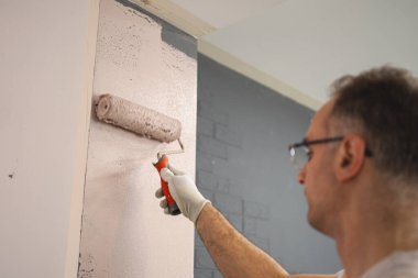 Duvarın önünde duran bir adam eşit boyayı uygulamak için bir silindir kullanıyor. Duvar her darbeyle şekil değiştiriyor. Adam yüzeyi yeni bir boya tabakasıyla kaplıyor..