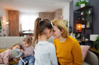 Anne en büyük kızıyla yakın temasta iken diğer iki kız kardeş oturma odasının arka planında koltukta oturuyor..