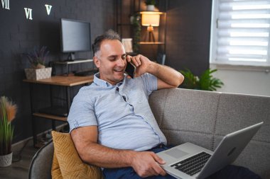 Kanepede dizüstü bilgisayar kullanırken telefonla konuşan orta yaşlı beyaz bir adam..