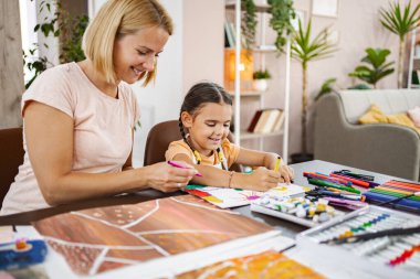 Bir anne ve küçük kızı sanat malzemeleriyle dolu rahat bir masada renkli çizimler ve yaratıcı aktiviteler yapıyorlar..