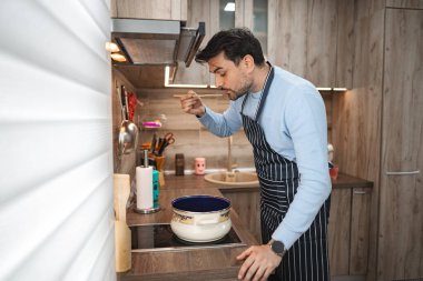 Adam mutfakta duruyor, tezgahta malzemeleri doğruyor ve pişiriyor. Etrafı çeşitli mutfak aletleri ve aletleriyle çevrili olarak işine odaklanmış durumda..