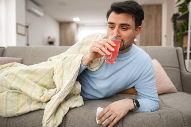 Battaniyeyle kaplı hasta adam oturma odasındaki kanepede uzanıyor, dinleniyor ve vitamin içiyor..