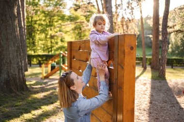 Gülümseyen anne, sevimli kızının oyun parkına çıkmasına yardım ediyor..