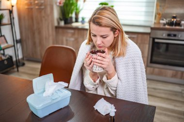 Genç bir kadın, mutfaktaki hastalıkla savaşırken sıcak bir içecekten yudumluyor..
