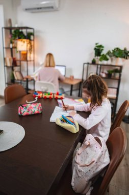 Ebeveyn yakınlardaki bir masada çalışırken çocuk ev ödevi yapar.