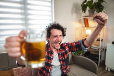 Genç bir adam televizyonda maç izlerken tezahürat yapıyor ve bira içiyor.