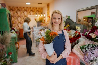 Mutlu bir çiçekçi, kendine has çiçek aranjmanlarıyla dolu canlı bir çiçekçide dururken bir buket renkli çiçek tutar. Arka planda çiçekleri düzenleyen başka bir çiçekçi görülebilir..
