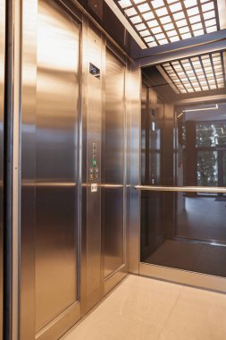 Şeffaf cam kapılı bir asansör açık duruyor ve önümüzde bir oda daha görünüyor. Asansörün mekanizması görünür, kablolar ve makaralar hareket halinde. Işık ışığı aydınlatır.