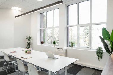 Beyaz bir masa ve sandalyeler, basit ve temiz bir estetik yaratacak şekilde bir odada düzgünce düzenlenir. Sandalyeler masanın etrafına yerleştirildi, kullanıma hazır. Oda doğal ışıkla ferah görünüyor.