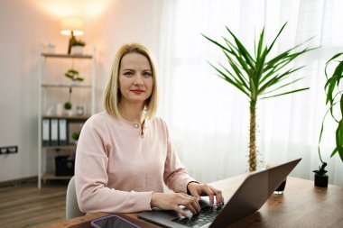 İyi ışıklandırılmış bir çalışma alanında dizüstü bilgisayarında omuz hizasında saçları olan bir kadın. Odada yeşil bir masa ve rahat bir masa var. Çalışırken odaklanmasını ve yaratıcılığını arttırıyor..