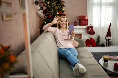 Noel ağacıyla süslenmiş şenlikli bir oturma odasında, gülümseyen bir kadın kanepesinde uzanır, akıllı telefonuna el sallar, arkadaşlarıyla veya ailesiyle keyifli bir video görüşmesi yapar..