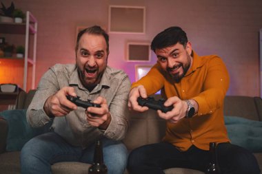 İki arkadaş heyecan verici bir oyun seansına daldılar, kontrolörler ellerinde, kahkahalar salona doluyor. Neşeli ifadeleri arkadaşça rekabetin neşesini yansıtıyor..