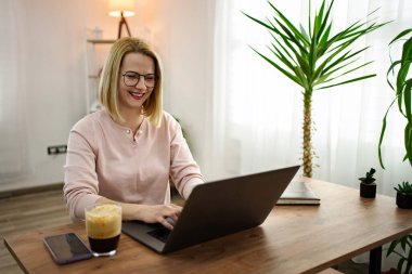 Bir kadın, uzak çalışma gününün tadını çıkarıyor. Ev ofisini aydınlık ve hoş karşılayan bir şekilde dizüstü bilgisayarında içki içerken daktilo ederek. Atmosfer bitkilerle ve yumuşak ışıklarla dolu..