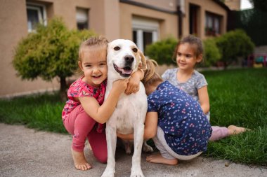 Üç neşeli çocuk yeşil bir bahçede neşeli bir köpeği kucaklıyor..