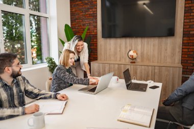 Farklı bireylerden oluşan bir grup, her biri dizüstü bilgisayar ekranlarıyla çevrili bir masanın etrafında oturmaktadır. Kağıtlar ve defterlerle işbirliği ya da işle ilgili bir faaliyete girmiş gibi görünüyorlar.