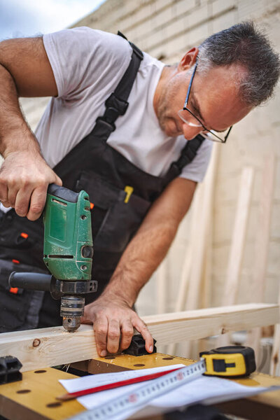 Зрелый плотник-мужчина с электрической отверткой на деревянной доске на открытом воздухе.