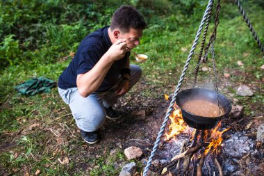 Genç adam ormanda yürüyüş gününde şenlik ateşinde pişirilen sıcak çorbayı tadıyor.