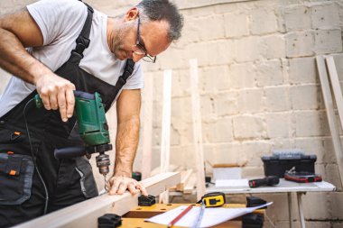 Olgun erkek marangoz, tahtadan bir tahta üzerinde elektrikli tornavida kullanıyor..