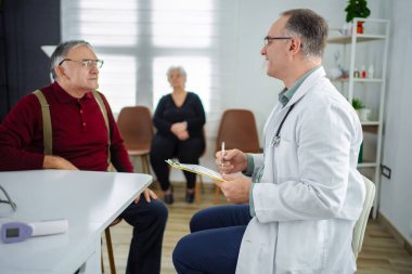 Doktor yaşlı bir adama danışırken, bir kadın yakınlarda gözcülük yapıyor..