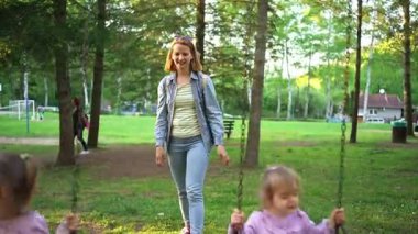 Genç bir kadın, salıncaklarda oynayan çocukların neşeli seslerine hayran bir şekilde yeşilliklerle dolu canlı bir parkta yürüyor. Atmosfer parlak ve neşeli, açık hava eğlencesi için mükemmel..