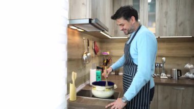 Sıcak bir mutfakta, bir adam, mutfak malzemeleri ve parlak dekor ile çevrili bir tencereyi zevkli bir yemek pişirme deneyimi içine daldırarak, mutlu bir şekilde karıştırır..