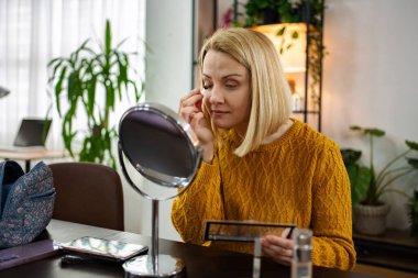 Sıcak ve davetkar bir mekanda, omuz hizasında sarı saçlı bir kadın aynaya bakarken makyajını dikkatlice yapar. Etrafında bitkiler ve kişisel eşyalar var. Huzuru kucaklıyor..