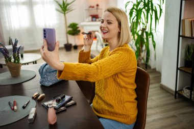 Neşeli bir kadın canlı bir videoyla meşgul oluyor, makyajını bitkilerle dolu sıcak dekore edilmiş bir yaşam alanında aynayla yapıyor. Güneş ışığı içeri akar ve parlak bir atmosfer yaratır..