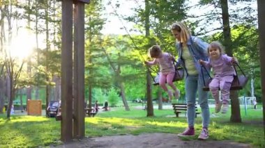 İki neşeli çocuk, neşeli bir şekilde güneşin altında sallanırken şefkatli bir yetişkin onları izliyor, canlı ağaçlarla çevrili ve huzurlu bir park düzeniyle..