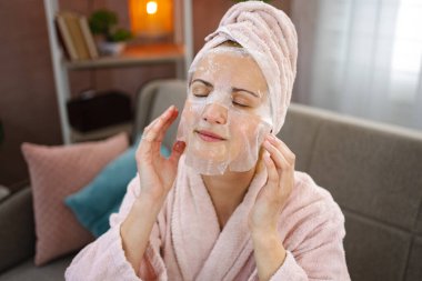 Bornoz ve havlu giymiş bir kadın oturma odasında nemlendirici kozmetik maske takıyor.