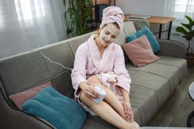 Kanepede oturan orta yaşlı bir kadın. Bornoz ve yüz maskesi takıyor. Evde taşınabilir lazer tüy temizleme cihazı kullanıyor.