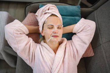 Kafasında bornoz ve havlu olan bir kadın, yüzünde nemlendirici bir maskeyle kanepede dinleniyor.