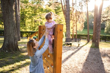 Gülümseyen anne, sevimli kızının oyun parkına çıkmasına yardım ediyor..
