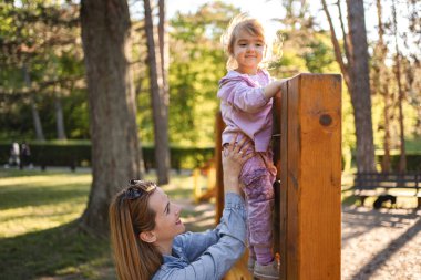Gülümseyen anne, sevimli kızının oyun parkına çıkmasına yardım ediyor..