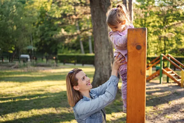 Gülümseyen anne, sevimli kızının oyun parkına çıkmasına yardım ediyor..