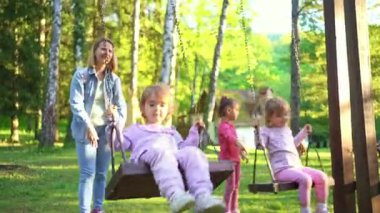 İki çocuk neşeli bir parkta mutlu bir şekilde sallanırken bir yetişkin gülümseyerek izliyor. Güneş ağaçların arasından süzülüyor, neşeli oyunlarını ve tasasız ruhlarını vurguluyor..