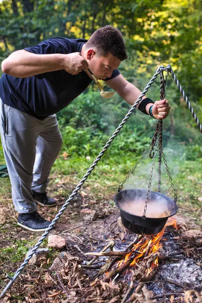 Genç kampçı, kamp ateşinde sallanan bir kazandan yemek tadıyor, vahşi bir yemek pişirme deneyiminin tadını çıkarıyor.