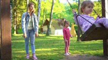 Çocuklar, bir yetişkinin eşliğinde salıncakta sallanmaktan zevk alırken kıkırdıyor. Güneş yeşil ağaçların arasından parlıyor, neşeli bir atmosfer yaratıyor..