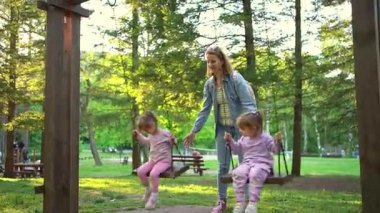 Canlı park atmosferi çocukların sallanırken kıkırdaması gibi havayı dolduruyor. Parlak yeşil bir günde anneleri onları sevgi ve kahkahalarla yüreklendiriyor..