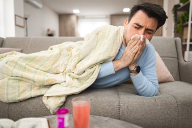 Adam bir kanepede uzanmış, yüzünü bir mendille örtüyor, hastalık ya da alerjiden dolayı burnunu siliyor. Gözleri sulu ya da yorgun görünebilir..