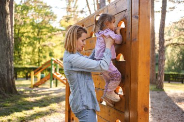 Gülümseyen anne, sevimli kızının oyun parkına çıkmasına yardım ediyor..