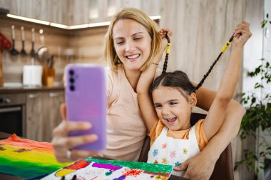 Bir anne ve kız, keyifli bir selfie anının tadını çıkarırken, aynı zamanda rahat mutfaklarında renkli çizimler ve el sanatları ile çevrili..