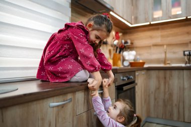 İki genç kız kardeş aralarındaki bağı rahat bir mutfak ortamında sergiliyorlar. Bir kız tezgahta neşeyle otururken, diğeri neşeyle onu aşağı çekmeye çalışarak hassas ve canlı bir atmosfer yaratmaya çalışıyor..