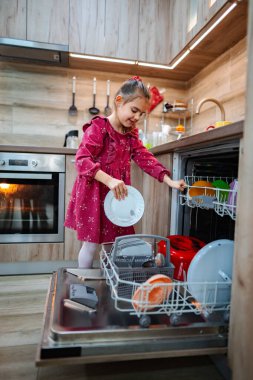 Parlak ve misafirperver bir mutfakta pembe elbiseli neşeli genç bir kız coşkulu bir şekilde bulaşıkları bulaşık makinesine yerleştiriyor. Sıcak güneş ışığı sıcak atmosferi güçlendirir..