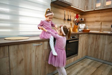 Konforlu ve şık bir mutfakta, biri tezgahın üzerine tünemiş, diğeri neşeyle uzanıp büyüleyici dünyalarında yaratıcılığı ve kahkahaları teşvik eden iki genç kız neşe dolu bir eğlenceye daldılar..