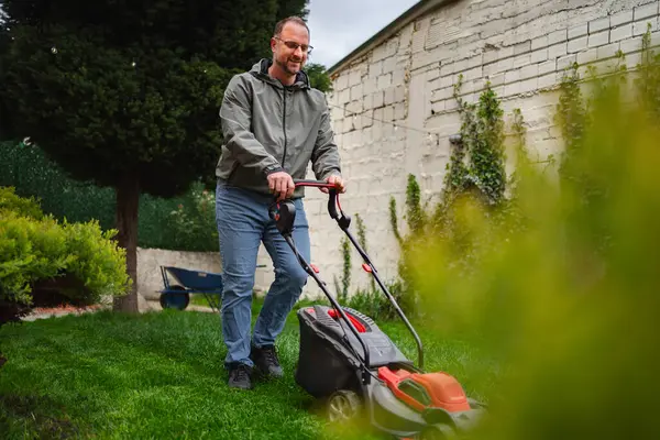 Neşeli bir adam yemyeşil çimlerini biçiyor, iyi korunmuş bir bahçede canlı bitkiler ve çalılarla çevrili. Gökyüzü açık, banliyöde keyifli bir öğleden sonrayı işaret ediyor..