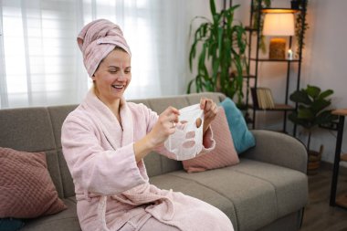 Kafasında bornoz ve havlu olan gülümseyen bir kadın, elinde bir yüz kozmetik maskesiyle oturma odasındaki kanepede oturuyor.