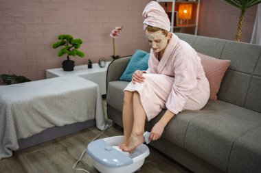 Kadın evinde elektrikli ayak banyosu yapıyor, maske takıyor ve bornoz giyiyor, rahatlamanın tadını çıkarıyor.